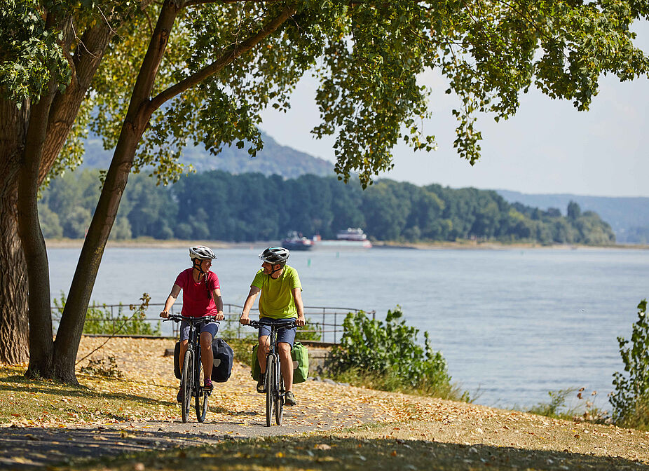 Fahrradausflug am Wasser Zwei Personen fahren Fahrrad, unterhalten sich, im Hintergrund ein Fluss.